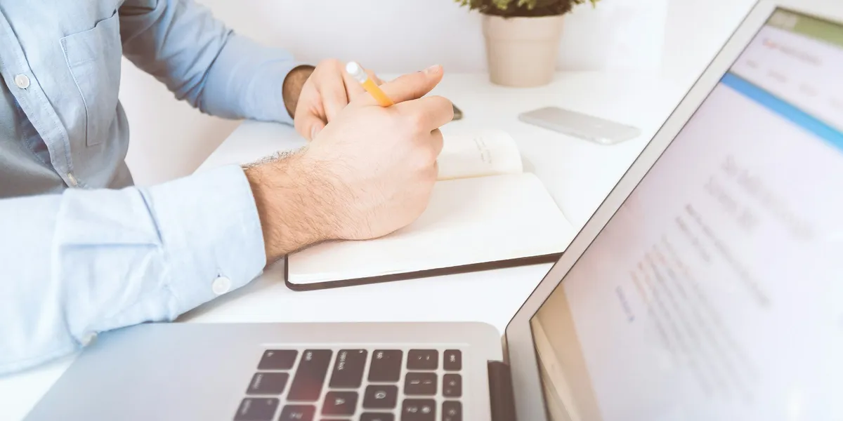 Mano che scrive su un quaderno accanto a un laptop aperto su una scrivania