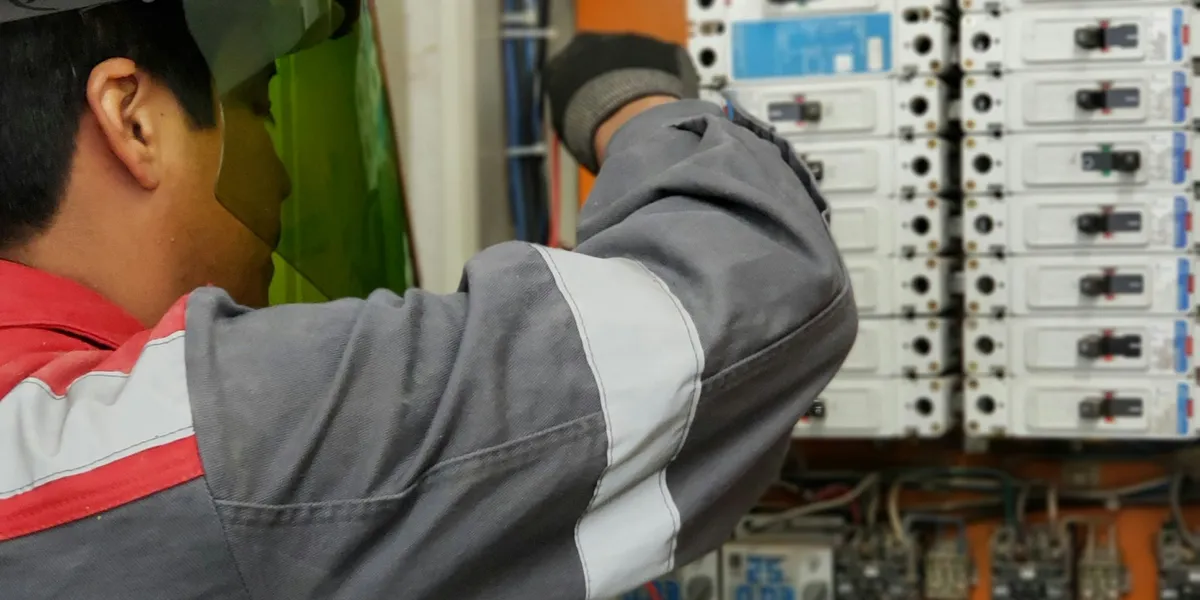 Tecnico con visiera protettiva e giacca da lavoro grigia e rossa interviene su un quadro elettrico aperto