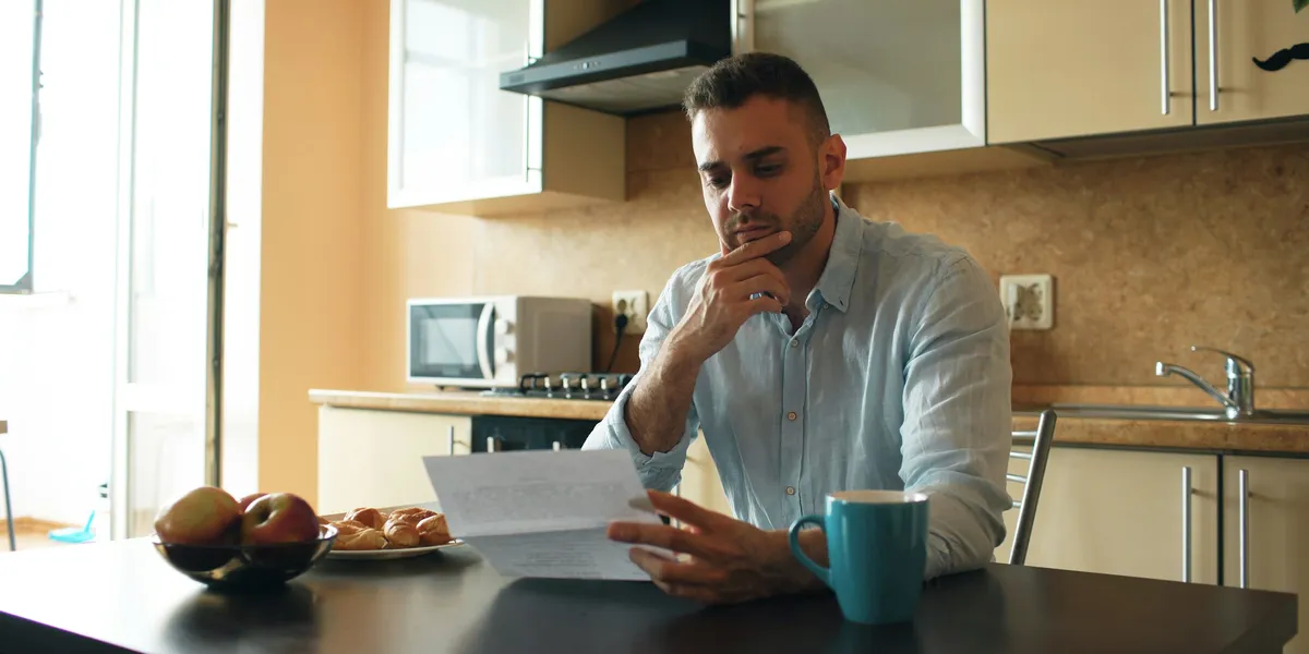 Uomo seduto al tavolo della cucina legge pensieroso un documento, con frutta, brioche e una tazza sul tavolo